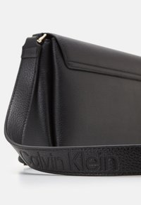 Svart läderhandväska med en strukturerad form, som har en texturerad yta och en präglad "Calvin Klein"-logotyp på remmen.