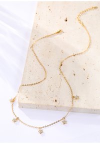 Złoty naszyjnik z łańcuszka z pięcioma charmsami w kształcie gwiazdek z przezroczystymi kamieniami szlachetnymi, na tle fakturowanego beżowego kamienia.