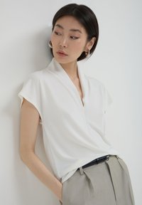 Donna con capelli corti e scuri che indossa orecchini a cerchio d'argento, una blusa avvolta bianca e pantaloni plissettati beige, appoggiata a un muro bianco semplice.