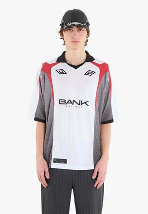 Giovane uomo con berretto nero, maglia sportiva bianca e rossa con la scritta "BANK HOLIDAY" e pantaloni scuri a righe sottili, in piedi davanti a uno sfondo bianco.