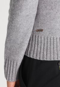 Grå stickad tröja med ribbad nederkant och en liten tygmärke med varumärke, som visar ett texturerat stickat mönster.