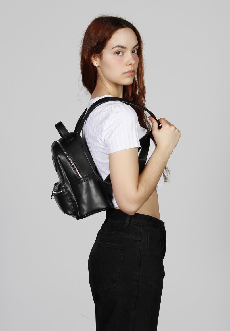 Mochila de cuero negro con forma redondeada, detalle de cremallera plateada y dos correas ajustables. El modelo lleva una camisa blanca corta y pantalones negros.