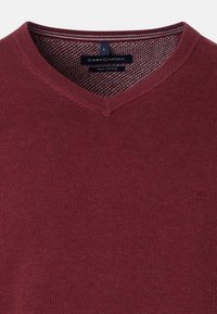Burgunderfarbener V-Ausschnitt-Pullover aus Pima-Baumwolle; mit geripptem Ausschnitt und Logodetail auf der unteren linken Seite. Glatte Textur.