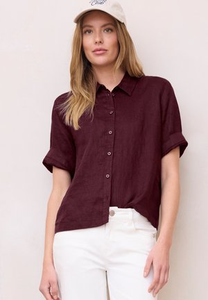 Nainen, jolla on beige lippalakki, tummanviininpunainen lyhythihainen nappipaita ja valkoiset housut, seisoo vaaleaa taustaa vasten.