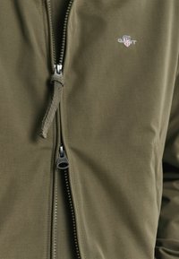 Green jacket made of smooth fabric with a full-length zip, toggle, and logo detail on the chest. Features a structured design.