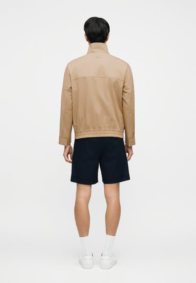 Veste légère beige avec un col montant, une taille élastique et deux poches latérales, associée à un short bleu marine et des baskets blanches.
