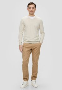 Hellebeige V-Ausschnitt-Pullover, cremefarbenes Unterhemd, khakifarbene Hose und weiße Sneaker. Einfaches Design ohne sichtbare Muster oder Akzente.
