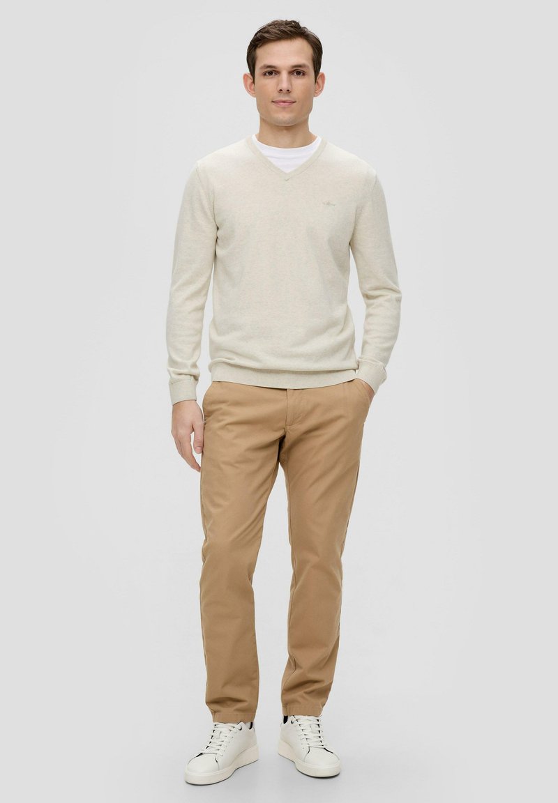 Hellebeige V-Ausschnitt-Pullover, cremefarbenes Unterhemd, khakifarbene Hose und weiße Sneaker. Einfaches Design ohne sichtbare Muster oder Akzente.