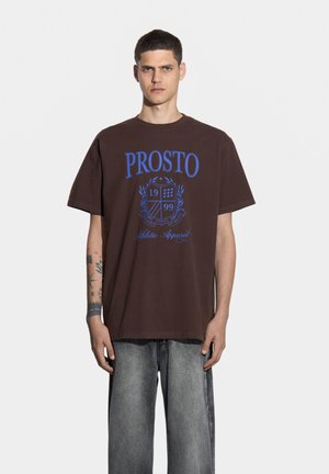 Brązowa koszulka bawełniana z niebieską grafiką "PROSTO", przedstawiająca wzór tarczy i rok "1999". Luźny krój i krótkie rękawy.