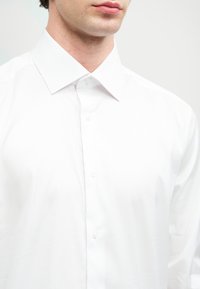 Man som bär en vit skjorta med knapp, med spridd krage, beskuren från näsan till mitten av torso, mot en ljus bakgrund.