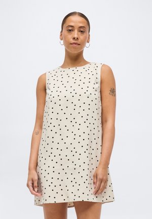 Donna con i capelli tirati indietro che indossa un vestito beige senza maniche con pois neri, in piedi contro uno sfondo bianco uniforme.