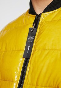 Giacca da uomo in nylon giallo lucido con collo a coste nero, dotata di chiusura a zip e di un'etichetta con marchio sulla cerniera.