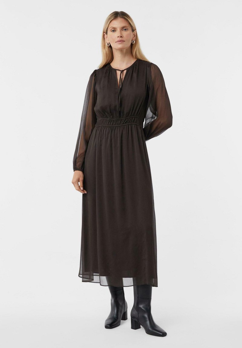 Schwarzes, langärmliges Kleid mit tailliertem Schnitt, leicht transparentem Stoff und gerafftem Design. Kombiniert mit knielangen schwarzen Stiefeln.