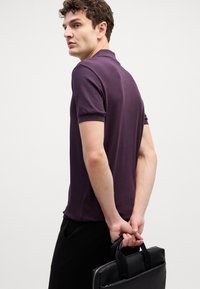 Tmavo purpurový polo tričko s krátkym rukávom, postava stojaca s čiernou koženou taškou, neutrálny pozadie, uvoľnený strih a hladká textura.