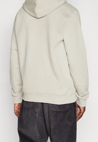 Osoba v béžové mikině a volných tmavých kordových kalhotách, zobrazená zezadu na světlém pozadí.