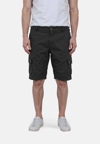 Mann in schwarzen Cargo-Shorts, weißen Sneakers und einem weißen T-Shirt, der mit entspannten Händen an den Seiten vor einem weißen Hintergrund steht.