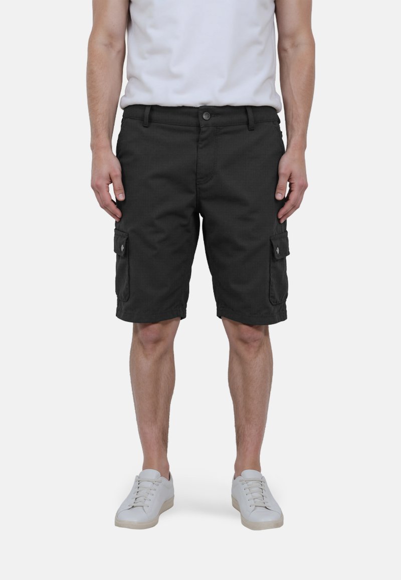 Mann in schwarzen Cargo-Shorts, weißen Sneakers und einem weißen T-Shirt, der mit entspannten Händen an den Seiten vor einem weißen Hintergrund steht.