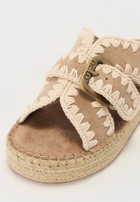 Sandale en suède beige avec deux brides croisées ornées de détails tissés contrastants et une boucle en laiton, montées sur une semelle en corde de jute.
