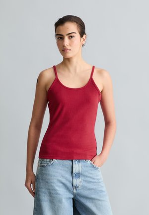 Jeune femme aux cheveux foncés portant un débardeur côtelé rouge et un jean denim bleu clair, debout devant un fond gris uni.
