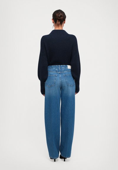 Vrouw met een knot in het haar, gekleed in een zwarte sweater, hoge taille, wijde blauwe jeans en zwarte hakken, met de rug naar een effen witte achtergrond.