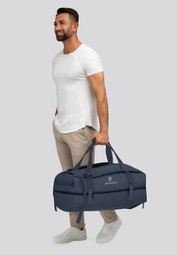 Sac de voyage bleu foncé avec doubles poignées, fermeture éclair et logo. Texture lisse et design sportif, porté par une personne en tenue légère.