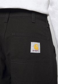 Schwarze Arbeitshosen aus strapazierfähigem Stoff, mit einer Gesäßtasche, die ein weißes Etikett mit einem gelben Logo und dem Text "carhartt" zeigt.