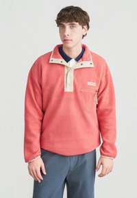 Νέος άνδρας που φοράει κορυφαίο φλις σε κοραλλί χρώμα με μπεζ πατιλέτα με κουμπιά και τσέπη στο στήθος με φερμουάρ, συνδυασμένο με μπλε παντελόνι, στέκεται μπροστά σε λευκό φόντο.