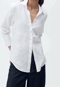 Chemise en lin blanc avec col cranté, manches longues et boutons à l'avant ; texture douce et coupe décontractée, associée à un pantalon en denim foncé.
