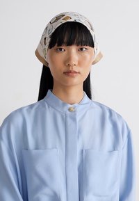 Mladá žena s hladkými čiernymi vlasmi, oblečená v modrej košeli na gombíky a vzorovanej šatke, s neutrálnym výrazom na tvári, na svetlošedého pozadí.