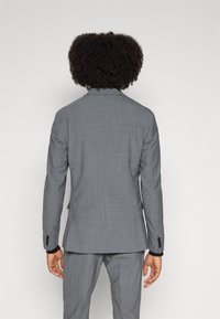 Persona con capelli ricci, che indossa un completo grigio e una camicia nera all'interno, in piedi di fronte a uno sfondo bianco uniforme.