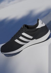 Sneaker Adidas bianca e nera con tre strisce bianche, suola ammortizzata e logo sul tallone, esposta su un piedistallo bianco con sfondo blu.