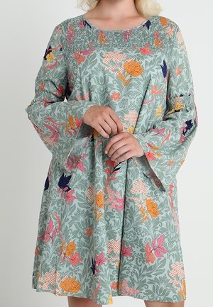 Femme portant une robe à manches longues vert clair avec des motifs floraux roses, orange et violets, et un détail smocké au niveau de l'encolure et des poignets.