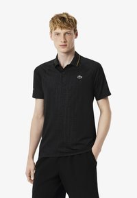 Svart polotröja med ett subtilt geometriskt mönster, gula accenter på kragen och en grön logotyp på vänster bröst. Tillverkad av slät tyg.