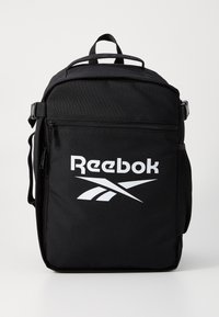 Črn Reebok nahrbtnik, izdelan iz trpežnega materiala, s prostornim žepom na zadrgo, ročajem na vrhu in izrazitim belim logotipom.