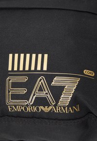 Tessuto nero con testo stampato in oro: "EA7 EMPORIO ARMANI" con un design a linee e accenti a strisce nella parte superiore. Superficie testurizzata.