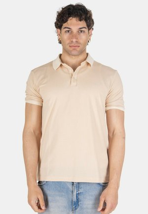 Giovane uomo con capelli scuri ricci, che indossa una polo beige a maniche corte e jeans azzurri, in piedi davanti a uno sfondo semplice.