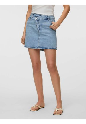 Femme portant une mini-jupe en jean bleu clair, un haut blanc sans manches et des sandales plates beiges à brides tressées, debout avec une main dans la poche.