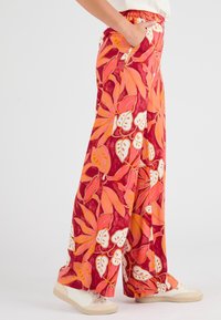Pantaloni rossi a gamba larga con un motivo floreale tutto intorno in arancione e bianco, dotati di una vita elastica e tasche. Abbinati a scarpe beige.
