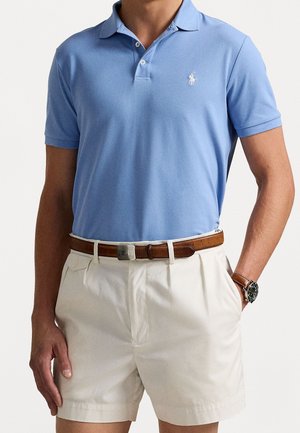 Polo i lyseblåt stof med klassisk krave og logo. Kombineret med cremefarvede shorts og et brunt læderbælte.