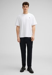 Witte katoenen t-shirt met korte mouwen, voorzien van een klein zwart bloemenlogo op de borst. Gecombineerd met zwarte broeken en zwarte schoenen.