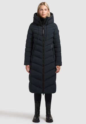 Abrigo de plumas largo y negro con un patrón en forma de chevron, que presenta un cuello alto, cierre con cremallera y una capucha. Llevado sobre leggings negros y botas.