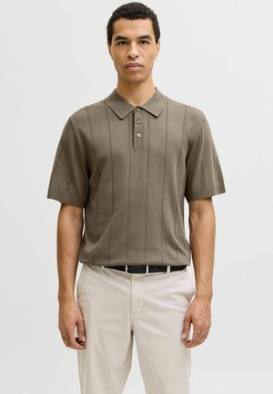 Mann som har på seg olivengrønn kortermet polotrøye med vertikale sømmer, beige bukser og svart belte, stående foran en ensfarget lys bakgrunn.