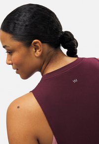 Top sin mangas de color marrón con un escote redondo, que muestra un logo visible en la parte posterior, combinado con una capa base de color claro por debajo.