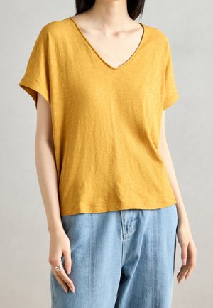 Kobieta ubrana w musztardowożółtą bluzkę z krótkim rękawem i dekoltem w serek oraz jasnoniebieskie dżinsy, stojąca na jednolitym szarym tle.