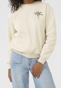 Sudadera color crema con cuello y puños acanalados, que presenta un gráfico de palmera verde en el pecho y un corte relajado. Jeans de color azul claro visibles.