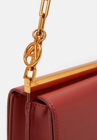 Borsa a tracolla in pelle rossa con una cornice in metallo dorato, catena e un emblema circolare. Presenta una forma strutturata e cuciture a contrasto.