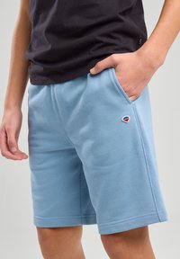 Un short bleu clair en tissu doux et texturé, doté de poches latérales et d'un petit logo brodé sur la jambe gauche.