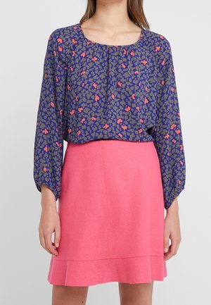 Vrouw die een lange mouwen blauwe blouse draagt met oranje en groene abstracte vlekken en een hoge taille roze rok met een geschulpte zoom.