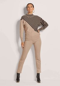 Beiger Pullover mit einem diagonalen schwarzen Streifenmuster auf einer Seite. Kombiniert mit figurbetonten beigen Hosen und schwarzen Ankle Boots.