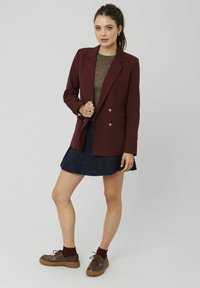 Blazer bordeaux à double boutonnage avec boutons dorés, associé à un haut scintillant et une jupe texturée bleu foncé, chaussures marron et chaussettes grenat.